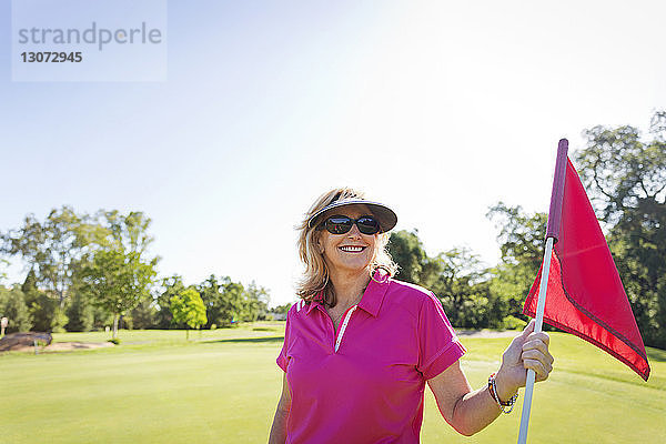 Porträt einer glücklichen Frau  die die Flagge hält  während sie am Golfplatz vor klarem Himmel steht
