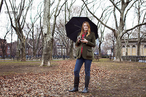 Frau mit Regenschirm schaut weg  während sie das Mobiltelefon im Park hält