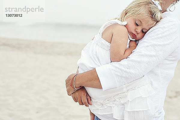 Mittelsektion einer Mutter  die ein Mädchen trägt  während sie am Strand steht