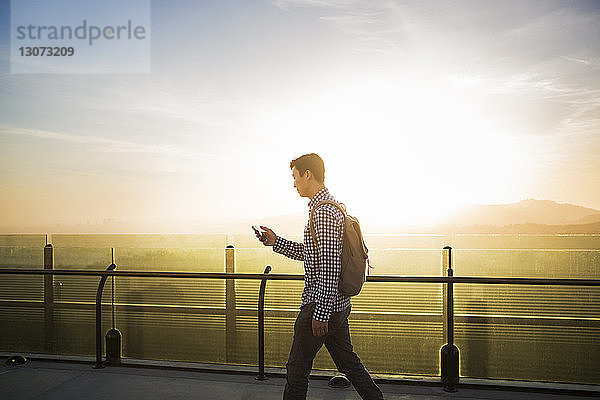 Seitenansicht eines Mannes  der beim Gehen am Geländer ein Smartphone benutzt