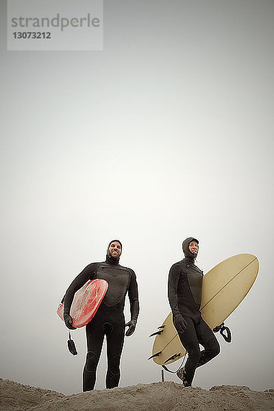 Freunde in Neoprenanzügen tragen Surfbretter  während sie gegen den klaren Himmel stehen