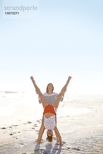 Porträt einer Frau mit den Beinen eines Freundes im Handstand auf Sand am Strand