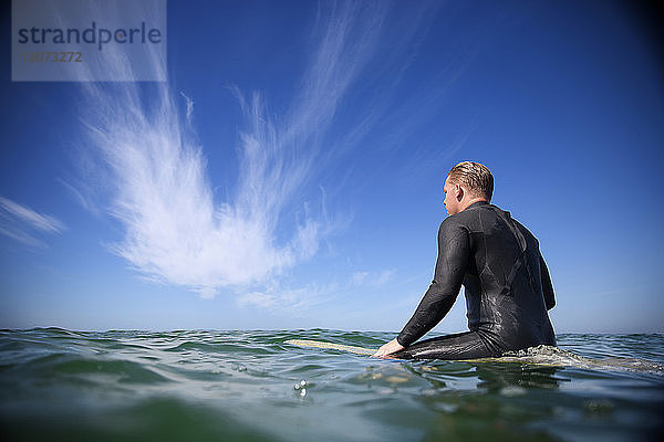 Rückansicht eines Mannes  der auf einem Surfbrett im Meer gegen den Himmel sitzt