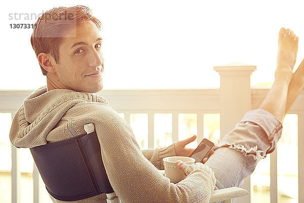 Porträt eines Mannes  der ein Smartphone in der Hand hält  während er auf dem Balkon sitzt