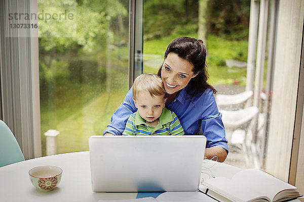 Glückliche Mutter sitzt mit ihrem Sohn zusammen  während sie den Laptop am Tisch gegen das Fenster benutzt