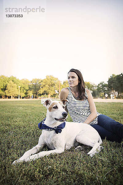 Nachdenkliche Frau sitzt mit Hund auf Grasfeld vor klarem Himmel im Park
