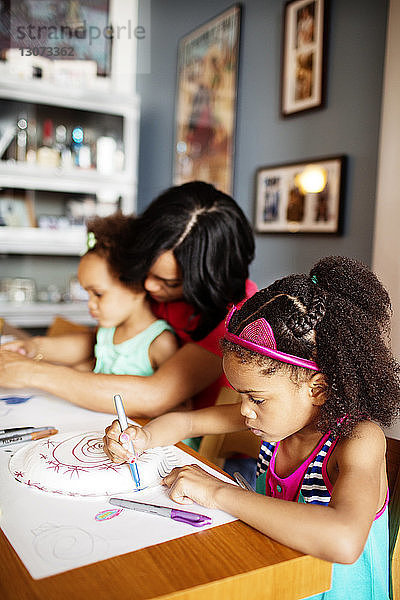 Mädchen  das auf Papier zeichnet  während es neben Mutter und Schwester am Tisch sitzt