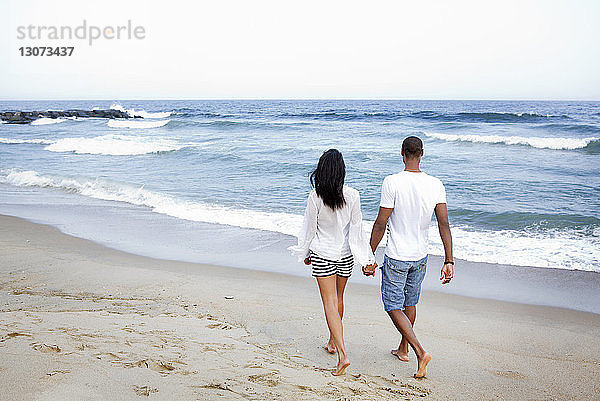 Rückansicht eines Paares  das sich beim Strandspaziergang am Ufer an den Händen hält