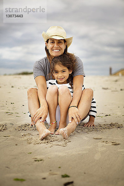 Porträt einer lächelnden Mutter und Tochter  die auf Sand vor wolkigem Himmel sitzen