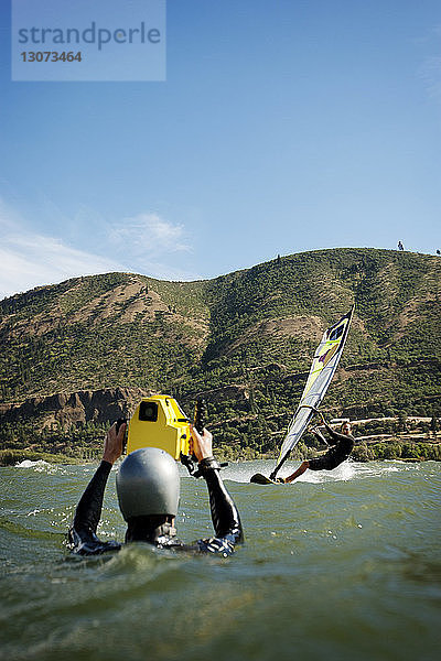 Mann fotografiert Freund beim Windsurfen im Meer gegen Berg