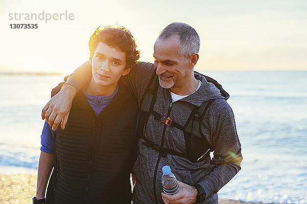 Porträt eines Sohnes mit dem Arm des Vaters um die Schulter gegen das Meer bei Sonnenuntergang