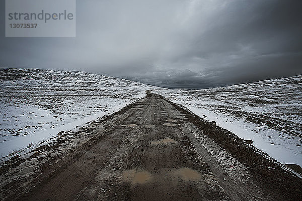 Unbefestigte Straße inmitten schneebedeckter Landschaft vor bewölktem Himmel