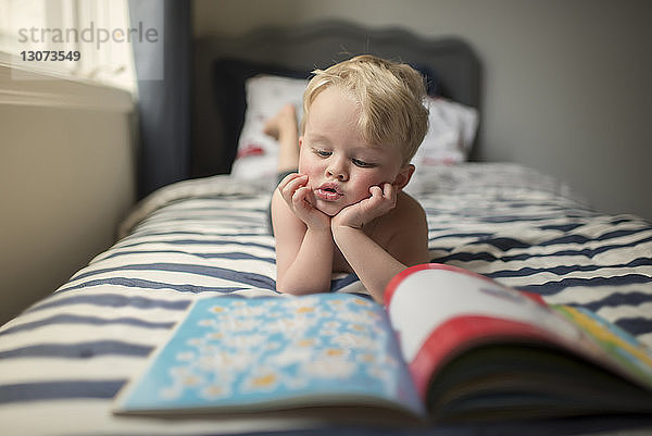 Junge mit Händen am Kinn liest Bilderbuch  während er zu Hause auf dem Bett liegt