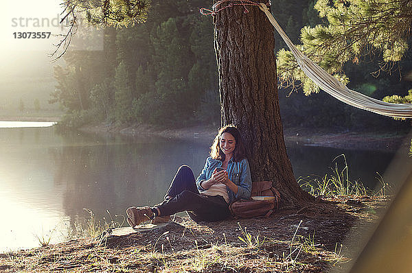 Glückliche Frau benutzt Mobiltelefon beim Entspannen am Seeufer