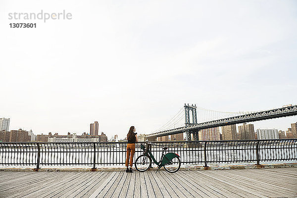 Frau mit Fahrrad steht auf Fußweg an der Brücke von Manhattan gegen den Himmel