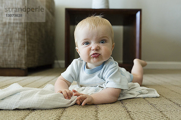 Porträt eines süßen  verspielten Jungen  der ein Gesicht macht  während er zu Hause auf dem Teppich liegt