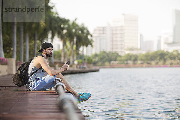 Nachdenklicher Mann  der mit Kopfhörern Musik hört und auf einem Holzsteg am Flussufer sitzt