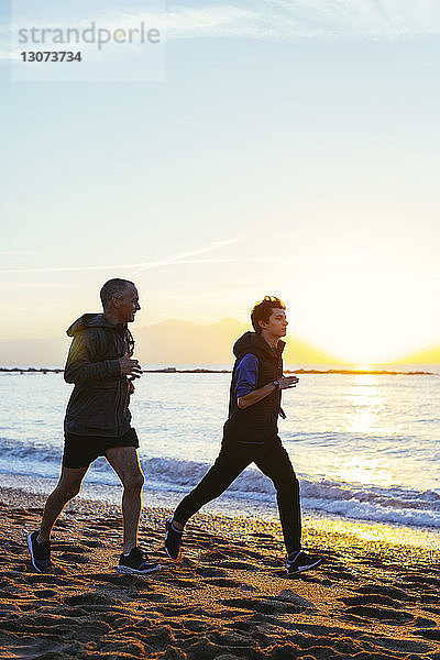 Zuversichtlicher Vater und Sohn joggen an der Küste am Strand gegen den Himmel bei Sonnenuntergang