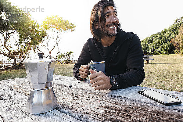 Glücklicher Mann mit Kaffee auf Holztisch  der weg schaut  während er gegen den Himmel sitzt
