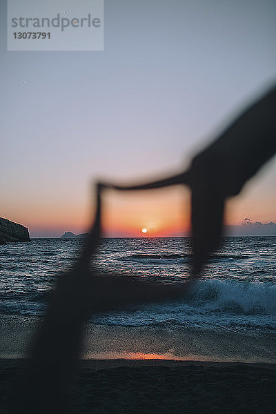 Sonne bei Sonnenuntergang durch den Rahmen der Hände gegen den Himmel am Strand gesehen