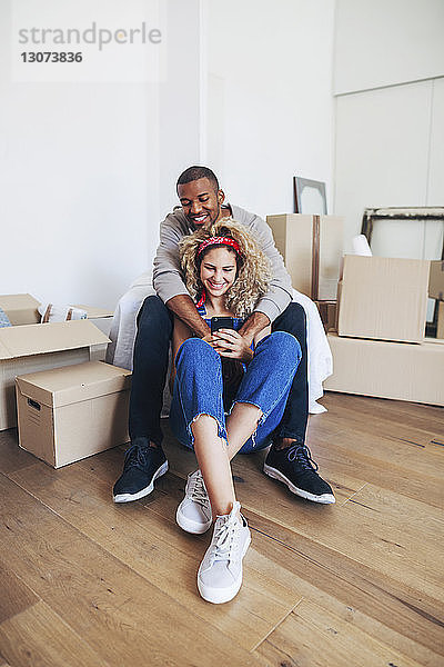 Glückliches Paar benutzt Mobiltelefon  während es in seinem neuen Zuhause sitzt