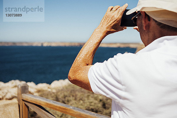 Rückansicht eines Mannes  der das Meer per Telefon fotografiert  während er auf einer Klippe steht