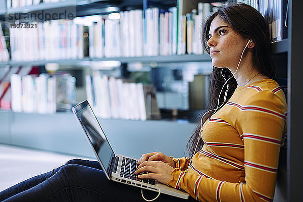 Nachdenkliche Frau mit Laptop-Computer sitzt an Bücherregalen in der Bibliothek