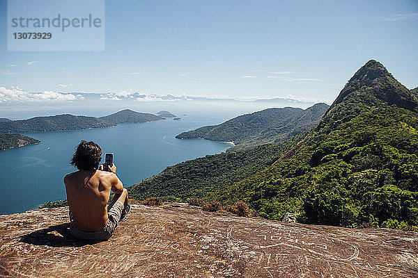 Rückansicht eines Mannes  der das Meer fotografiert  während er auf einem Berg gegen den Himmel sitzt
