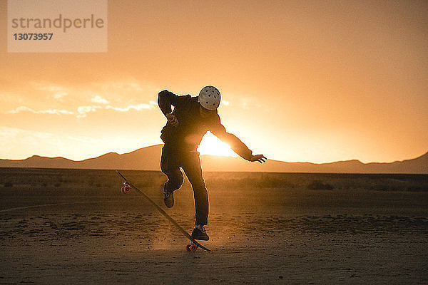Mann in voller Länge beim Stunt mit Skateboard auf dem Feld gegen den Himmel bei Sonnenuntergang