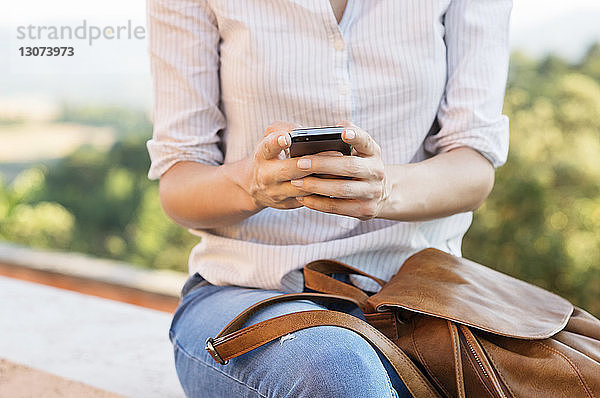 Mittelsektion einer Frau  die ein Smartphone benutzt  während sie auf der Stützmauer einer Terrasse sitzt