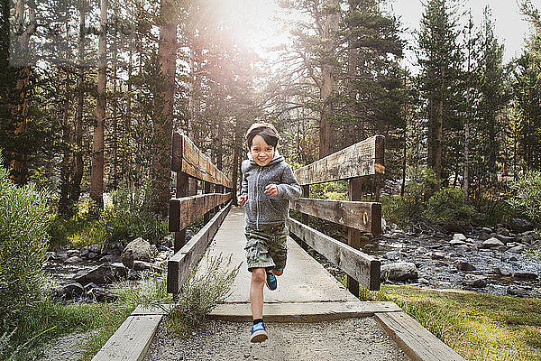 Porträt eines Jungen  der im Wald über eine Holzbrücke gegen Bäume rennt