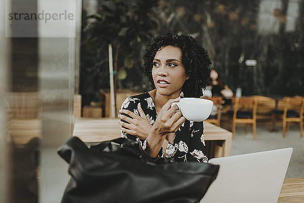 Nachdenkliche Frau hält Kaffeetasse in der Hand  während sie im Café am Laptop-Computer sitzt