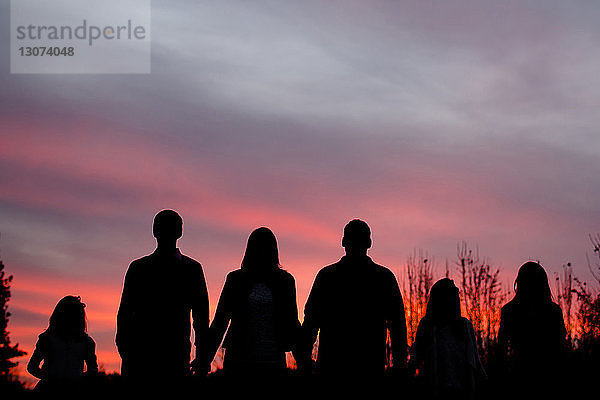 Scherenschnittfamilie hält sich an den Händen und steht bei Sonnenuntergang vor dramatischem Himmel auf dem Feld