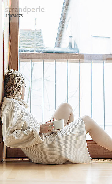 Seitenansicht einer Frau mit Kaffeetasse  die durch das Fenster schaut  während sie zu Hause auf dem Boden sitzt