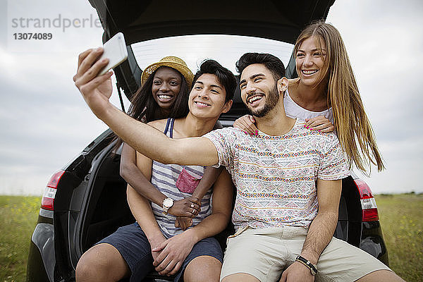 Glückliche multiethnische Freunde  die sich im Kofferraum eines Autos auf dem Spielfeld sitzend per Smartphone selbstständig machen