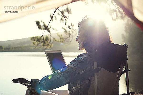 Lächelnde Frau mit Tablet-Computer  die sich bei Sonnenschein auf einem Sessel am Seeufer entspannt