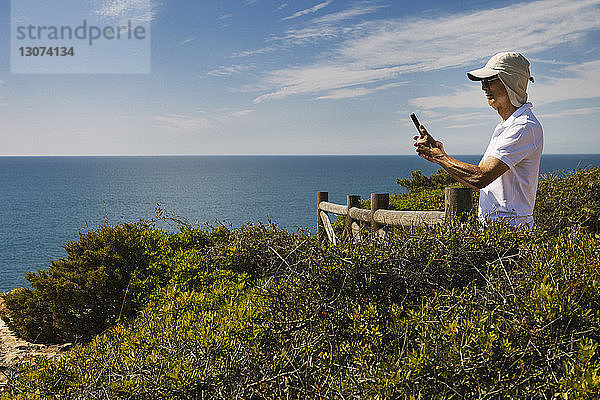 Mann benutzt Telefon  während er auf einer Klippe am Meer gegen den Himmel steht