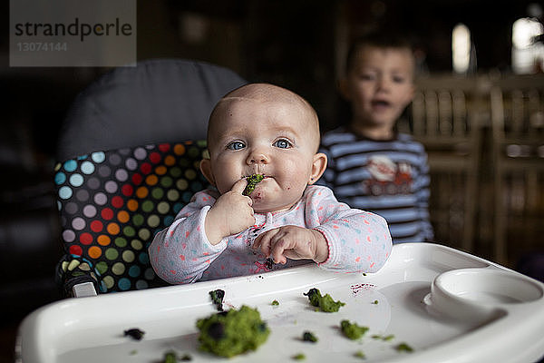 Kleines Mädchen isst Essen auf Hochstuhl  während der Bruder im Hintergrund steht