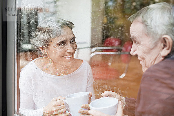 Glückliches älteres Ehepaar unterhält sich  während es Kaffeetassen in der Hand hält  die durch ein Glasfenster gesehen werden