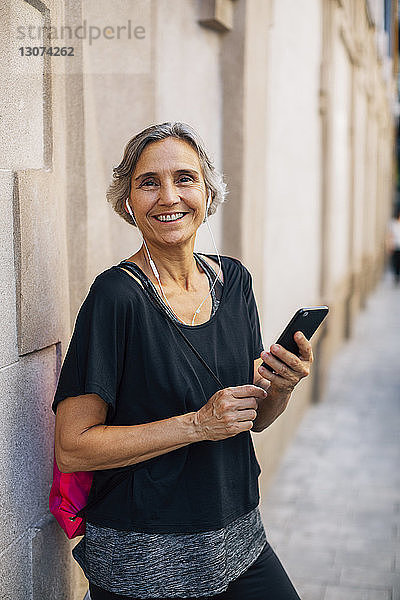 Porträt einer glücklichen Frau  die ein Smartphone in der Hand hält  während sie an der Wand steht