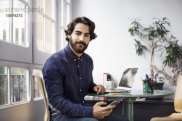 Porträt eines selbstbewussten Geschäftsmannes  der ein Smartphone in der Hand hält  während er im Büro sitzt
