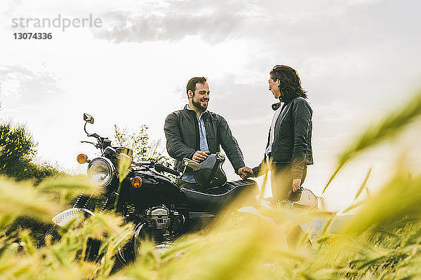 Ehepaar redet  während es mit dem Motorrad auf einem Grasfeld gegen den Himmel steht