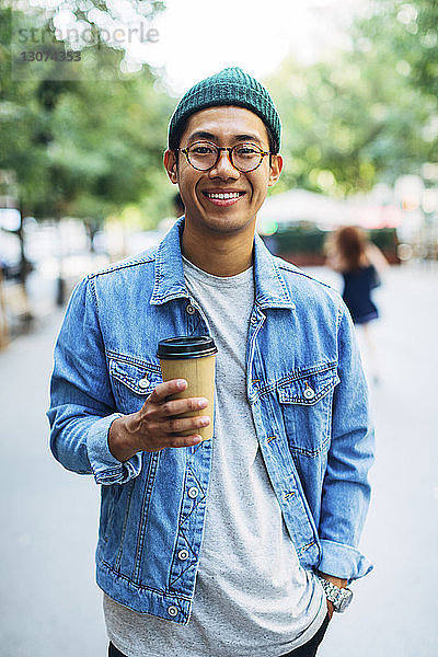Porträt eines glücklichen Mannes mit der Hand in der Tasche  der einen Einwegbecher am Bürgersteig hält