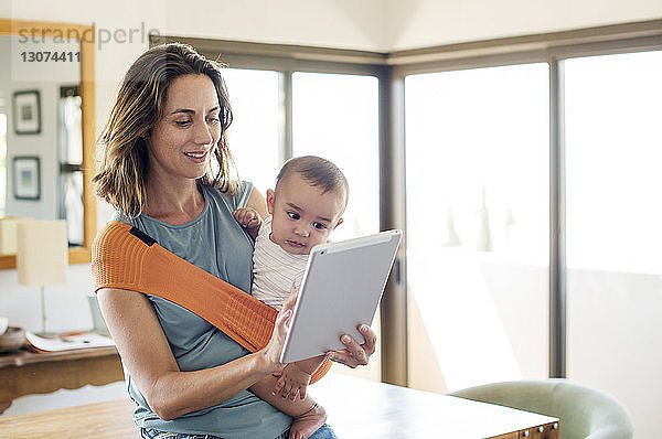 Mutter  die einen kleinen Jungen in einer Babytrage trägt  während sie zu Hause einen Tablet-Computer benutzt