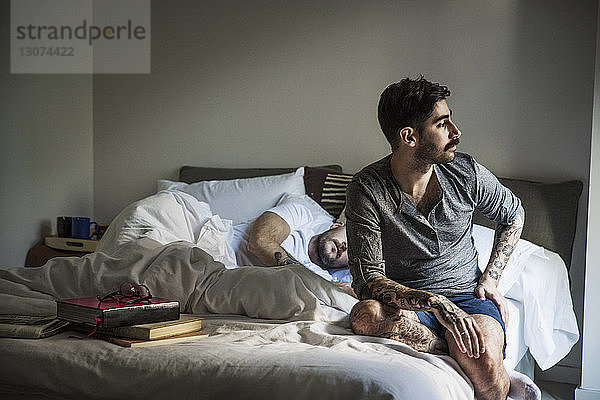 Nachdenklicher schwuler Mann schaut weg  während der Partner auf dem Bett schläft