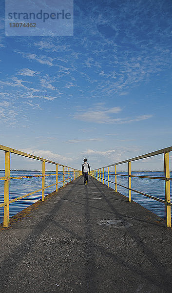 Rückansicht eines Jungen  der auf dem Pier über den Fluss geht  bei bewölktem Himmel