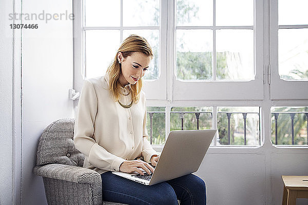 Lächelnde Geschäftsfrau benutzt Laptop  während sie im Büro auf einem Stuhl sitzt
