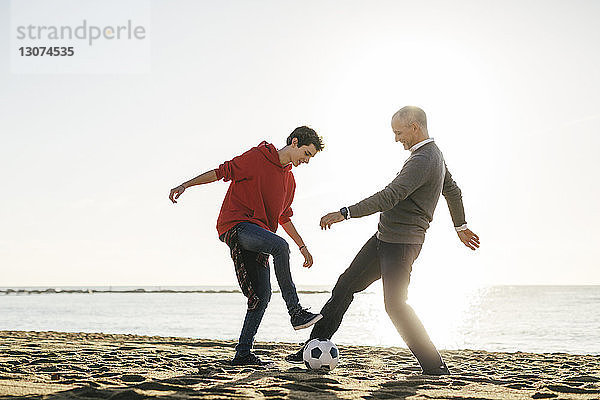 Vater und Sohn in voller Länge beim Fußballspielen am Strand vor klarem Himmel