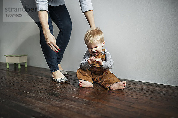 Niedrige Sektion der Mutter steht neben dem fröhlichen kleinen Jungen  der zu Hause auf dem Boden sitzt