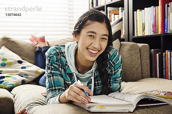 Porträt einer fröhlichen Teenagerin  die Hausaufgaben macht  während sie zu Hause auf der Couch liegt